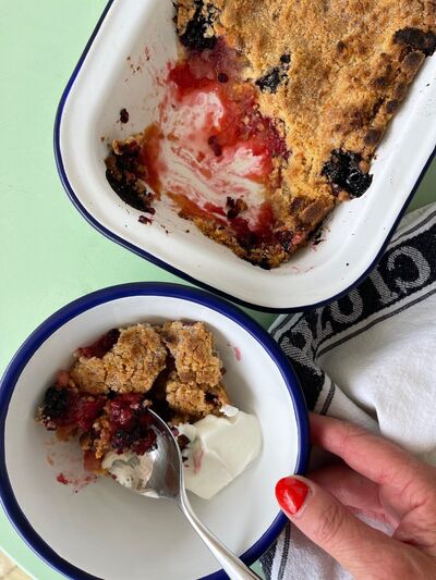 Blackberry and apple crumble in a white enamel dish, with a bowl served with cream and a spoon