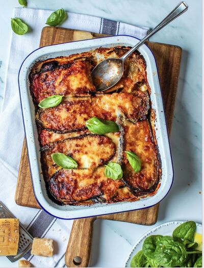 A layered aubergine and tomato bake topped with melted cheese and basil in a white enamel dish