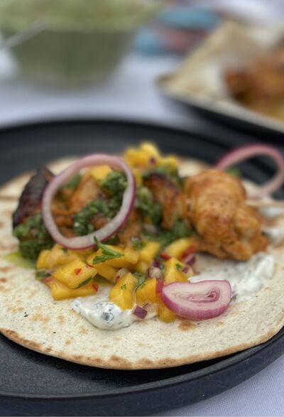 Chicken kebab on flatbread with mango salsa, green chutney, red onion and yoghurt, on a dark plate