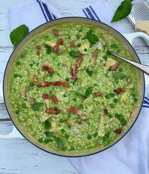 A pan of green pea risotto topped with crispy bacon pieces, basil leaves and grated parmesan