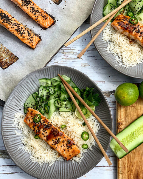 A bowl of glazed salmon on rice with sliced cucumber and green chilli, with chopsticks and limes beside it