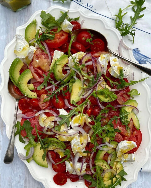 A platter of tomatoes, avocado, red onion, rocket and pieces of mozzarella with serving spoons