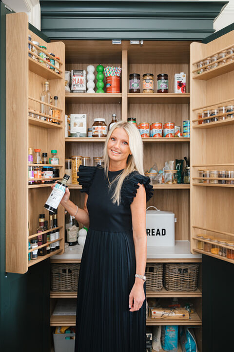 Mandy in a navy dress smiling and holding a bottle at an open oak pantry full of jars, tins and baskets