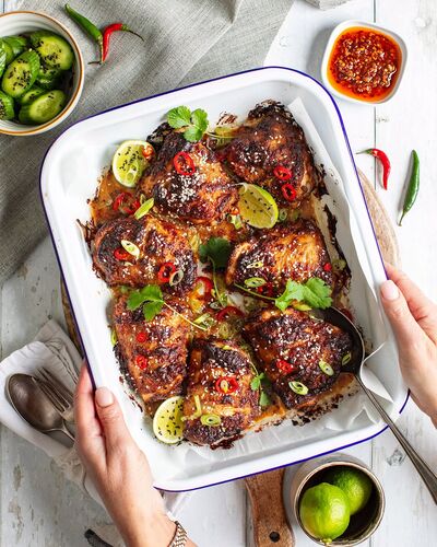 Glazed chicken thighs sprinkled with sesame seeds, sliced red chilli, spring onion and coriander, with lime wedges, in a white enamel tray held in two hands, with a bowl of sliced cucumber and a dish of chilli sauce beside it