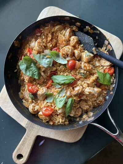 A black skillet of sausage and tomato orzo with cherry tomatoes and torn basil, on a wooden board