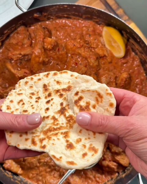 Hands holding a torn, charred flatbread over a pan of rich orange-red chicken curry with half a lemon in the sauce