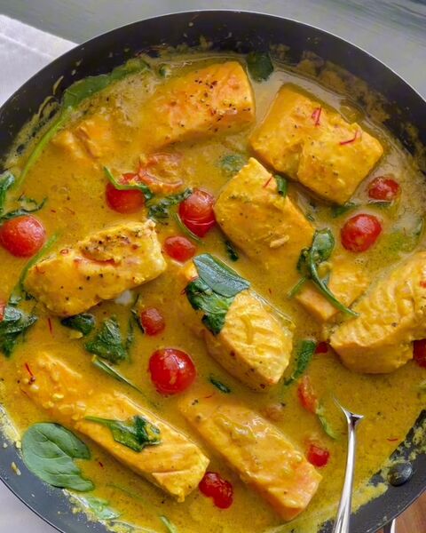 Pieces of salmon in a golden-yellow sauce with spinach and cherry tomatoes, in a dark pan on a wooden board